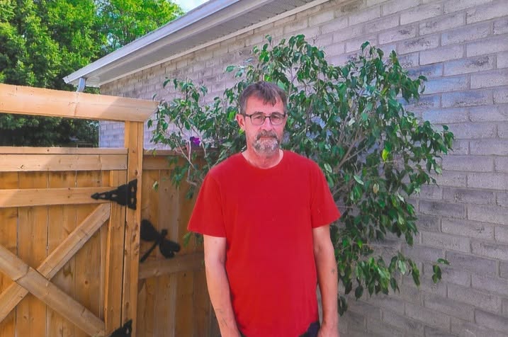 Kevin standing outdoors beside a wooden gate, wearing a red shirt